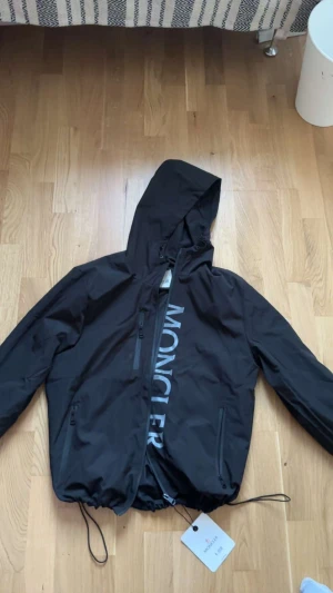 Svart vindjacka från Moncler - Svart vindjacka från Moncler med stor logotyp på framsidan och broderad patch på ärmen. Jackan har huva, dragsko i nederkant och två fickor med dragkedja. Materialet är lätt och syntetiskt, perfekt för blåsiga dagar. Snygg och stilren design med Moncler-tag kvar. Nfc tag gär inte att scannas