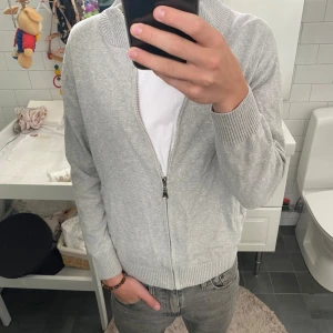 Ljusgrå stickad kofta med dragkedja - Snygg ljusgrå stickad kofta med hel dragkedja och ribbad krage. Perfekt för lager-på-lager och funkar till både jeans och t-shirt. Enkel och stilren design som passar till det mesta.