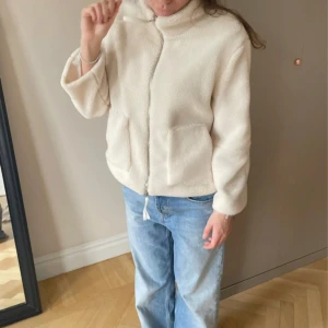 Beige teddyjacka med dragkedja - Mysig beige teddyjacka med hög krage och två stora fickor framtill. Jackan har en dragkedja med snörning i nederkant och är långärmad med loose passform. Perfekt för dig som gillar chill och comfy stil.