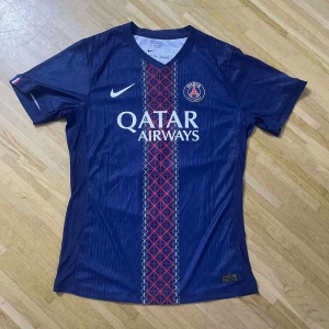 PSG Barcola 29 matchtröja Nike - Säljer en officiell Paris Saint-Germain fotbollströja med Barcola och nummer 29 på ryggen. Tröjan är marinblå med rött och vitt grafiskt mönster längs mitten, PSG-logga och Qatar Airways tryck på bröstet. Kortärmad och tillverkad i lätt, ventilerande material.