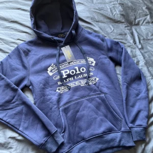 Marinblå hoodie från Polo Ralph Lauren. Prisförlsag! - Snygg marinblå hoodie från Polo Ralph Lauren med vit tryckt logga på bröstet. Tröjan har huva med snörning och en stor magficka framtill. Mjuk och skön bomullsmix, perfekt för chill dagar. Klassisk streetstil med ribbade muddar vid ärmar och nederkant.