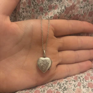 Silverfärgat hjärthalsband med medaljong - Säljer ett fint halsband med hjärtformat medaljonghänge i silverfärg. Medaljongen har graverade detaljer och går att öppna för att lägga in en liten bild eller minne. Kedjan är tunn och elegant, perfekt för att ge din stil en personlig touch. 