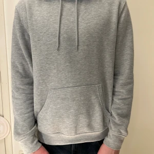 Grå hoodie med magficka och huva - En stilren grå hoodie med klassisk magficka och snörning i huvan. Tröjan har lång ärm och normal passform, perfekt för chill dagar. Materialet är mjukt och känns skönt mot huden. Enkel design utan mönster eller logga, passar dig som gillar clean look.