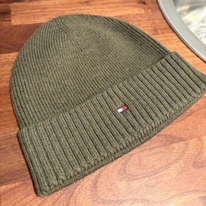 Mössa från Tommy Hilfiger - Snygg olivgrön mössa från Tommy Hilfiger med ribbad design och uppvikt kant. Perfekt för dig som vill ha en stilren och enkel look under kalla vinterdagar. Skicket är nyskick då den aldrig är använd.