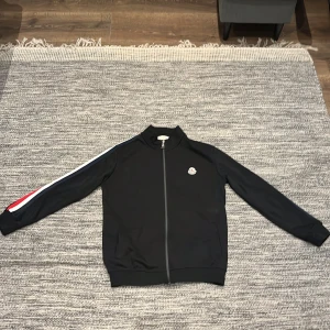 Svart Moncler kofta M - Svart kofta från Moncler med dragkedja framtill och broderad logga på bröstet. Koftan har långärmade armar med röd och vit. Tillverkad i mjuk fleece för en skön och sportig vibe.