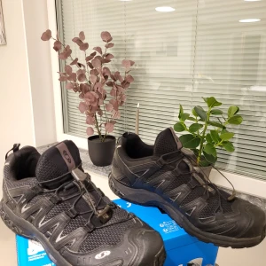 Salomon XA 3D Ultra 2 svarta sneakers - Salomon XA 3D Ultra 2 sneakers i svart med grå detaljer. Skorna har meshpaneler för bra ventilation, robust syntetmaterial och snabbsnörning. Perfekta för äventyr och outdoor med slitstark sula och sportig design. Märket syns tydligt på sidan och plösen.