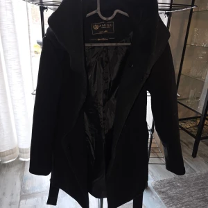 Svart kappa med bälte från Amisu, så gott som ny - Stilren svart kappa från Amisu med snygg krage och bälte i midjan. Jackan har långa ärmar och är fodrad på insidan för extra komfort. Perfekt för dig som vill ha en klassisk look med modern touch.