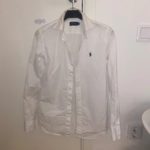 Vit skjorta från Polo Ralph Lauren - Klassisk vit skjorta från Polo Ralph Lauren med blå broderad logga på bröstet. Skjortan har lång ärm, knappar framtill och är tillverkad i bomull. Perfekt för en clean och stilren look.