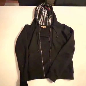 Svart Burberry hoodie  - Svart hoodie från Burberry med klassisk rutig huva i svart, vit och röd. Tröjan har snörning vid huvan och långärmade ärmar. Perfekt för dig som vill ha en stilren och lyxig look med ikoniskt Burberry-mönster. Kan diskutera pris!