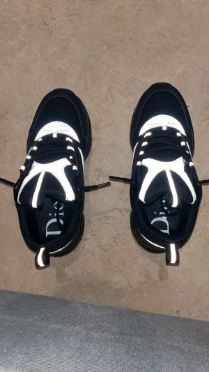 Svarta Dior sneakers med reflexdetaljer - Chunky svarta Dior sneakers med silvriga och vita reflexdetaljer. Ovandel i mesh och skinn, med kraftig sula och snörning. Snygg sportig vibe och tydlig Dior-logga på sidan. Perfekta för dig som gillar statement-sneakers med modern design.