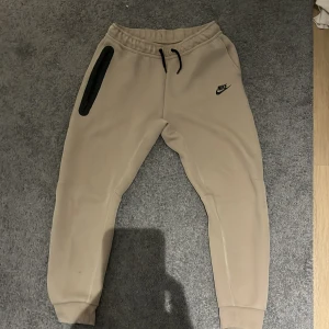 Beige mjukisbyxor från Nike XL - Snygga beige mjukisbyxor från Nike i storlek XL, med svart Nike-logga och svart snörning i midjan. Byxorna har en cool dragkedja på sidan och ribbade muddar vid bensluten. Perfekta för chill eller träning, tillverkade i mjukt bomullsmaterial. Med litet hål bak till