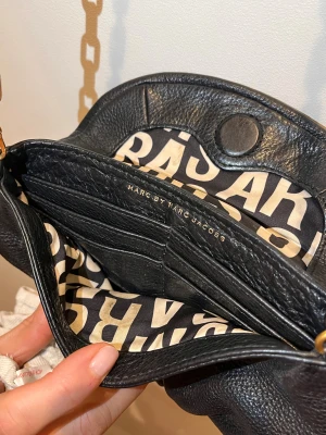 Svart axelväska Marc by Marc Jacobs - Snygg svart axelväska från Marc by Marc Jacobs i äkta skinn med guldfärgade metalldetaljer och kedja. Väskan har ett coolt innerfoder med grafiskt mönster och en ikonisk metallplatta framtill. Perfekt storlek för det viktigaste och riktigt trendig look.