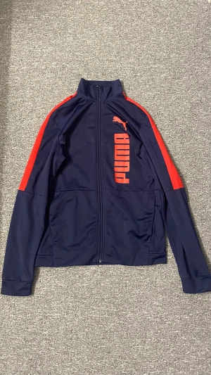 Blå och röd Puma vindjacka - Snygg vindjacka från Puma i mörkblått med röda detaljer längs axlarna och ett stort rött Puma-tryck på framsidan. Jackan har hel dragkedja, hög krage och långa ärmar. Perfekt för dig som gillar sportig stil och vill sticka ut med färg.
