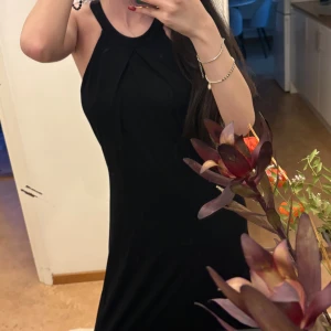 Svart långklänning med halterneck - Svart långklänning med snygg halterneck och stilren design. Klänningen har en elegant detalj framtill med veckad tyg och en knapp i nacken. Passformen är figurnära och materialet känns mjukt mot huden. Perfekt för dig som gillar minimalistisk och chic stil.