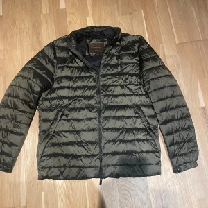 Mörkgrön pufferjacka från Massimo Dutti - Snygg mörkgrön pufferjacka från Massimo Dutti med horisontella quiltade sömmar och dragkedja framtill. Jackan har hög krage, två sidofickor och långa ärmar med knappdetalj vid mudden. Perfekt för kyliga dagar och har en clean, modern look.