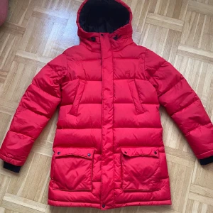Dunjacka - Säljer en röd puffer-/dunjacka från Everest med stor huva och svarta detaljer. Jackan har två stora fickor med lock och knappar, samt två snedställda bröstfickor. Fodret är svart med mönster och tillverkat med Ecoya-teknologi. Perfekt för kalla dagar! Butikspris för ny är ca 1500kr. Priset kan diskuteras.