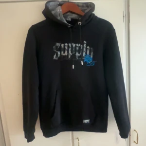 Svart hoodie Supply & Demand XS - Svart hoodie från Supply & Demand med camouflagemönstrad huva och snörning. Tryck på bröstet med texten 'supply' och en blå grafisk detalj. Mjuk insida och känguruficka framtill. Perfekt för dig som gillar streetwear och coola detaljer.