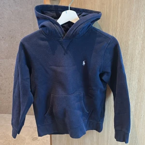 Marinblå hoodie från Polo Ralph Lauren - Snygg marinblå hoodie från Polo Ralph Lauren med klassisk logga broderad på bröstet. Tröjan har huva, magficka och långa ärmar. Lite urtvättat runt magen.