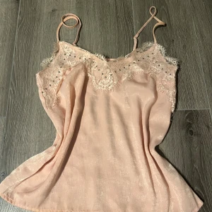Ljusrosa linnetopp med spetsdetaljer - Supersöt ljusrosa linnetopp med tunna axelband och vacker spets längs halsringningen. Toppen har små silverfärgade pärlor i spetsen och är tillverkad i ett glansigt, mjukt material som ger en lyxig känsla. Perfekt för dig som gillar romantiska och feminina vibes.