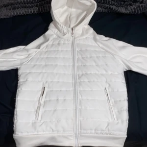 Vit quiltad pufferjacka med huva - Säljer en fräsch vit pufferjacka med quiltad framsida och huva. Jackan har dragkedja hela vägen och två fickor med dragkedja. Perfekt för dig som gillar stilren och clean look. Materialet känns mjukt och lätt, och passformen är normal.