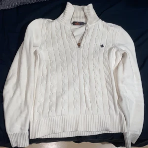 Vit kabelstickad tröja Morris S - Kabelstickad vit tröja från Morris med halv dragkedja och hög krage. Tröjan har ribbade muddar och en broderad logga på bröstet. Perfekt för lager-på-lager och snygg till jeans eller kjol.