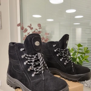 Svarta mockakängor Chriss stl 39 - Svarta kängor från Chriss i mocka med grov sula och inbyggda broddar för extra grepp. Fodrade med mjukt material för värme och komfort. Snygga snördetaljer och robust design, perfekta för kalla dagar och hala underlag.