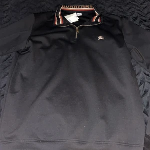 Svart halvzip tröja från Burberry - Svart långärmad tröja från Burberry med halv dragkedja och klassisk logga på bröstet. Kragen har Burberrys signaturmönster och broderad text. Tillverkad i 100% bomull för en mjuk och bekväm känsla.