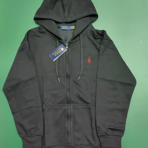 Svart hoodie från Polo Ralph Lauren - Svart hoodie från Polo Ralph Lauren med röd broderad logga på bröstet. Tröjan har huva med snörning, dragkedja framtill och ribbade muddar. Perfekt för dig som gillar klassisk och stilren streetwear.
