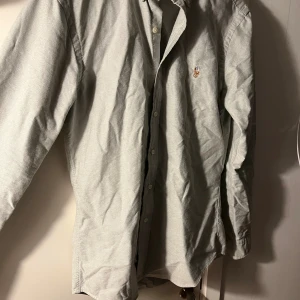 Grå skjorta från Polo Ralph Lauren - Slim fit skjorta i ljusgrått från Polo Ralph Lauren med klassisk krage och broderad logga på bröstet. Skjortan har långa ärmar och knappar framtill. Materialet är mjuk bomull som känns skönt mot huden och passar perfekt till en clean och stilren look. Skriv frågor och bättre bilder kan lösas