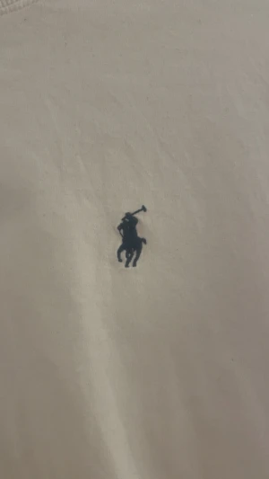 Vit t-shirt från Polo Ralph Lauren - Vit t-shirt från Polo Ralph Lauren med klassisk rund hals och korta ärmar. T-shirten är i mjuk bomull och har den ikoniska broderade loggan i marinblått på bröstet. Enkel och stilren design som funkar till allt.