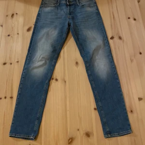 Blå jeans från Jack & Jones - Säljer ett par klassiska blå jeans från Jack & Jones med straight fit. Jeansen har snygga slitningar framtill och bakfickor med diskret söm. De är tillverkade i jeansmaterial och har normal passform med knapp och dragkedja i midjan. Storlek 33/36