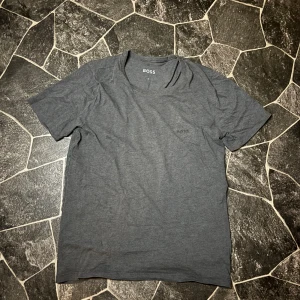 Hugo Boss Tshirt  - Snygg grå t-shirt från BOSS med diskret broderad logga på bröstet. Modellen är klassisk med rund hals och korta ärmar, perfekt för en clean och enkel stil. Materialet är mjukt och skönt bomullstyg som känns bekvämt hela dagen.