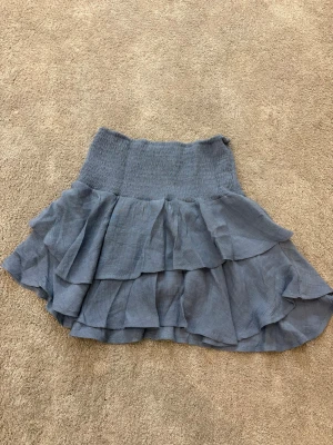 Blå volangkjol med resårmidja - Supersöt blå kortkjol med flera lager volanger och bred resår i midjan. Kjolen har en luftig och flowy känsla, perfekt för att skapa en chill och trendig look. Materialet känns lätt och skönt mot huden, och färgen är en snygg dov blå som passar till mycket.