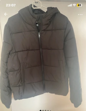 Svart pufferjacka från Pieces - Svart pufferjacka från Pieces med huva. Jackan har en grov dragkedja framtill, två sidofickor. Perfekt för kalla dagar och har en clean, enkel look som funkar till de flesta stilar. Har ett litet hål nere vid sömmen inne i jackan. Inget som syns på. 