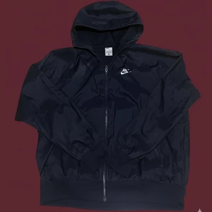 Svart vindjacka från Nike med huva - Svart vindjacka från Nike med klassisk logga på bröstet. Jackan har huva med snörning, hel dragkedja framtill och ribbade muddar. Tillverkad i lätt polyester som passar perfekt när du vill ha en sportig och clean look