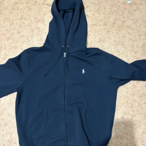 Marinblå zip- hoodie från Polo Ralph Lauren - Säljer en marinblå hoodie från Polo Ralph Lauren med dragkedja och huva. Tröjan har snörning vid huvan, två fickor fram och den klassiska Polo-loggan broderad på bröstet. Perfekt för chill dagar och enkel att matcha med jeans eller joggers.