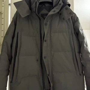 Grå dunjacka från Canada Goose M - Säljer en grå dunjacka från Canada Goose med snygg hög krage och huva. Jackan har dragkedja och knappar framtill, samt två vertikala fickor med dragkedja. Märkets patch syns på ärmen och insidan är fodrad med polyester. Perfekt för kalla dagar.