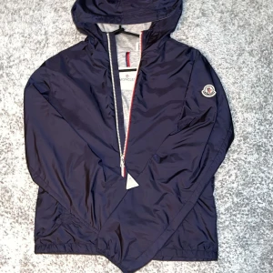 Mörkblå vindjacka från Moncler - SKRIV FÖR FRÅGOR.    Mörkblå Moncler Vindjacka – Stil, Komfort och Lyx i EttSnygg mörkblå vindjacka från Moncler med huva och klassisk logga på ärmen. Jackan har en rak passform, hel dragkedja med röd och blå detalj, samt elastiska muddar vid midja och ärmslut. Perfekt för dig som vill ha en stilren och sportig look.