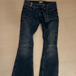 Mörkblå bootcut jeans med slitsar - Snygga mörkblå jeans med bootcut passform och slitna detaljer på benen. Jeansen har klassiska fem fickor, bälteshällor och en knytning i midjan. Perfekt för dig som gillar en avslappnad och trendig stil.