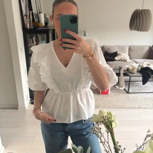 Vit blus med volang från Pernilla Wahlgren - Supersöt vit blus från Pernilla Wahlgren med v-ringning, puffiga trekvartsärmar och breda volanger med broderade detaljer längs axlar och bröst. Blusen har resår i midjan för en snygg passform och är gjord i mjuk bomull. Perfekt för dig som gillar romantisk och feminin stil.