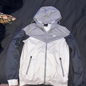 Nike vindjacka XS grå & vit - Nike vindjacka i storlek XS med huva och dragkedja framtill. Jackan har en snygg tvåfärgad design i ljusgrått och vitt, samt praktiska fickor med dragkedja. Skicket är 7,5/10. Nypris 1,700, skicka ett meddelande vid fler frågor