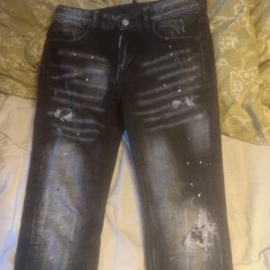 Svarta Dsquared2 slim jeans (passar nån som är 172cm) - Snygga svarta slim jeans från Dsquared2 med coola slitningar och färgstänk för en edgy look. Jeansen har klassisk femficksdesign, tydliga slitdetaljer och en tvättad finish. Märkespatch bak och Dsquared2-logga vid gylfen. Perfekta för dig som gillar streetstyle.