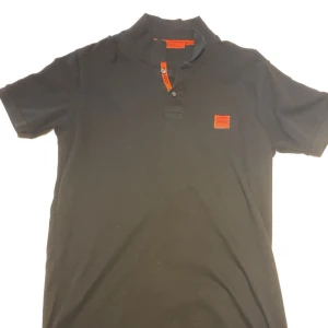 Svart Hugo Boss t-shirt med röd logga - Svart t-shirt från Hugo Boss i storlek XL med klassisk krage och knappar framtill. Snygg röd Hugo-logga på bröstet och röda detaljer vid knäppningen. Tillverkad i mjuk bomull för en skön känsla. Perfekt för en stilren och modern look.