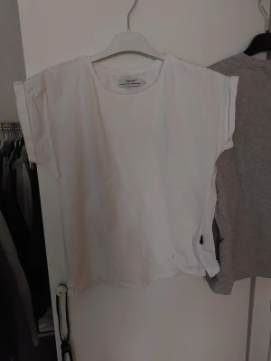Vit topp från Dedicated, oanvänd - Säljer en enkel och clean vit topp från Dedicated. Toppen har rund halsringning och ärmlös design med breda axlar. Tillverkad i mjuk bomull som känns skön mot huden. Perfekt för dig som gillar minimalistisk stil och vill ha en basic topp i garderoben.