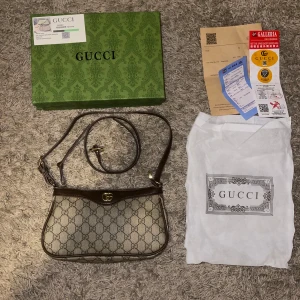 Gucci axelväska med GG-mönster - Snygg axelväska från Gucci i beige canvas med klassiskt GG-mönster och bruna skinn-detaljer. Väskan har justerbar axelrem, guldfärgad logga och dragkedja upptill. Kommer med originalbox och dustbag. Perfekt för dig som vill ha en ikonisk designerbag.