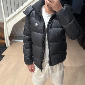 Vinterjacka Calvin Klein - Svart pufferjacka från Calvin Klein med CK-logga på bröstet. Jackan har huva, dragkedja framtill och två sidofickor med dragkedja. Den är quiltad och har en loose passform, perfekt för kalla dagar. Materialet är syntet och jackan är långärmad. Köpt från NLY MAN för ca ≈2300kr