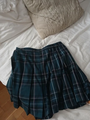 Mörkgrön rutig plisserad kjol H&M - Snygg mörkgrön plisserad kjol från H&M med klassiskt rutigt mönster i vitt och rött. Kjolen har en kort längd och är perfekt för dig som gillar preppy stil. Materialet känns som bomull och polyester, vilket gör den bekväm och lätt att bära. Passar dig som vill sticka ut med en cool look.