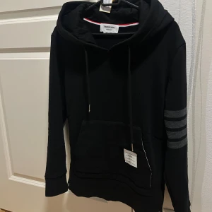 Thom Browne. Hoodie - Svart hoodie från Thom Browne med huva och snörning, samt klassiska grå ränder på ena ärmen. Tröjan har en stor magficka och detaljer med röd, vit och blå rand på insidan av ficka och ärmslut. Tillverkad i mjuk bomull, perfekt för chill dagar.