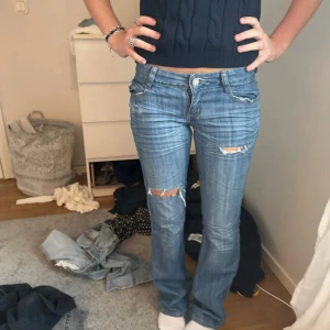 Blå bootcut jeans med broderade fickor unika - Snygga blå jeans med bootcut passform och slitna detaljer på knäna. Baktill har byxorna broderade fickor med text och mönster, samt en liten läderpatch i midjan. Jeansen har låg midja och klassisk femficksdesign. Perfekt för dig som gillar en cool och avslappnad stil.