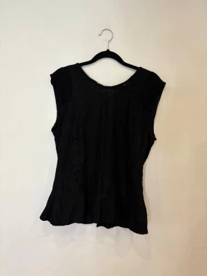 Svart spetsblus med volangkant - Säljer en svart blus med spetsdetaljer och volangkant nertill. Blusen är ärmlös och har en rund halsringning. Perfekt för dig som gillar en stilren men ändå romantisk look. Materialet är mjukt och följsamt med vacker spets över hela framsidan.